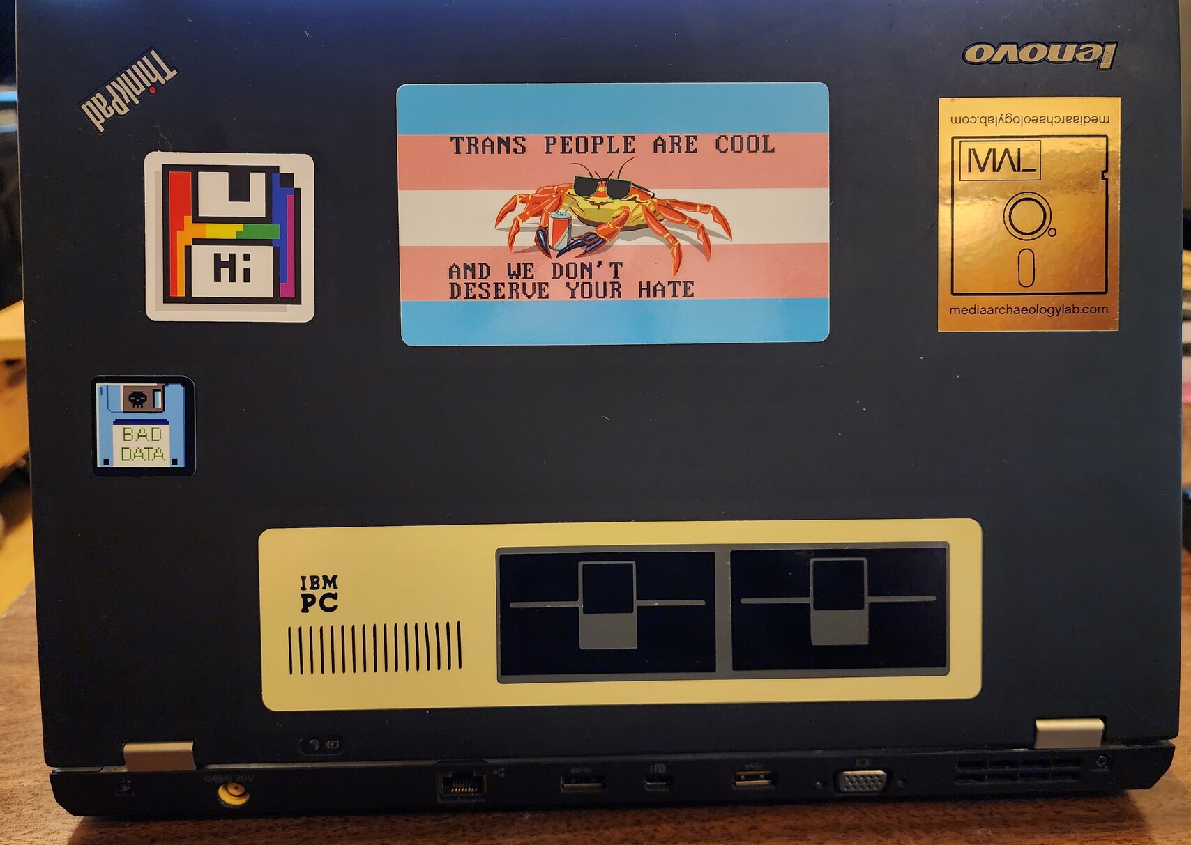 Back of a laptop. There's a trans pride flag sticker with a cool crab on it, and the text "trans people are cool, and we don't deserve your hate"

There's also three floppy disk stickers and an IBM PC sticker 
