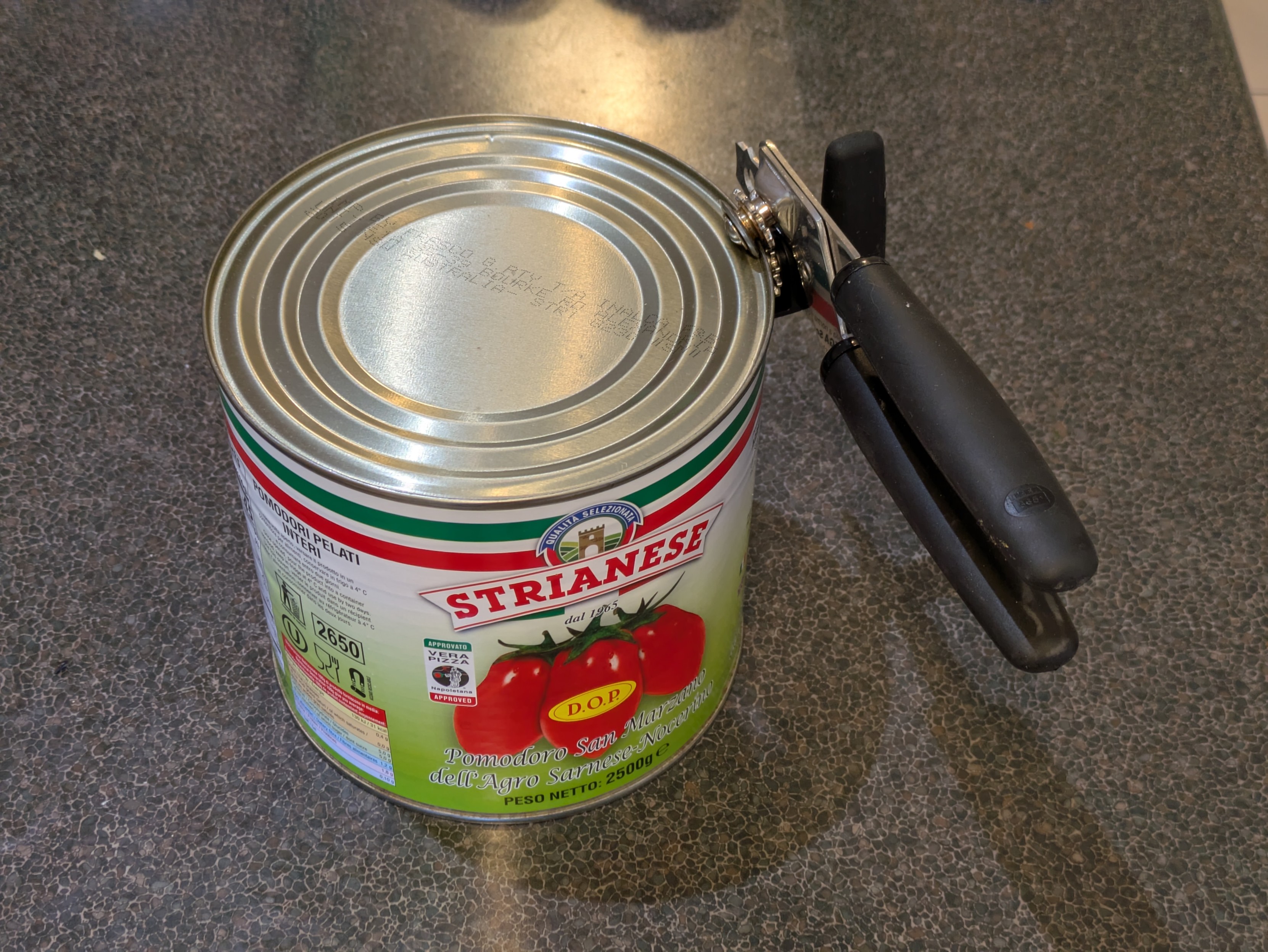 A photo of a huge 2.5kg can of San Marzano tomatoes from Italy sits on a home kitchen bench. A rotary can opener hangs off the top edge of the can, as if ready for action.